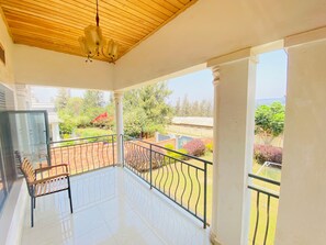 A balcony with a wooden ceiling, a bench, and a view of a garden with trees and a building in the background.
