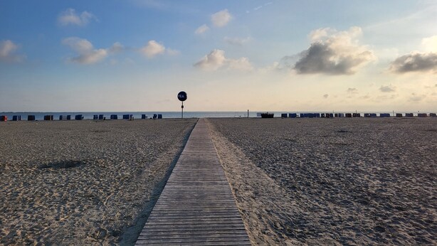A beach with a wooden walkway leading to the sea, a sign with 'SEA' on it, and a row of beach huts in the background. - Haus Seelöwe für 3 Personen, Strandnah, Terrasse, Grill, Fahrräder, Wlan, top (Dornum)