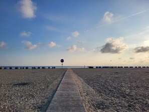 Una playa con un pasillo de madera que lleva al mar, un cartel con la palabra 'SEA' y una hilera de cabañas de playa al fondo.