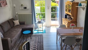 A living room with a sofa, a coffee table, a rug, a dining table with chairs, a bookshelf, and a window with curtains. - Haus Seelöwe für 3 Personen, Strandnah, Terrasse, Grill, Fahrräder, Wlan, top (Dornum)