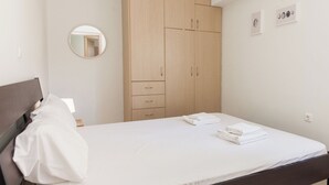 A neatly made bed with white linens, a wooden wardrobe with multiple drawers, a round mirror on the wall, and a small framed picture collection. - Comfy New Apt w Balcony GC 3360-01 (Athens)