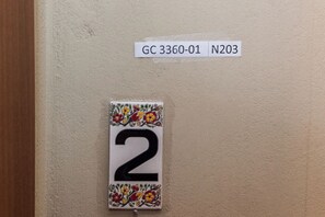 A room number sign with a floral border, a door, and a wall-mounted label with room identifiers. - Comfy New Apt w Balcony GC 3360-01 (Athens)