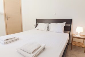 A neatly made bed with white linens and pillows, a wooden nightstand with a lamp, and a closed door in the background. - Comfy New Apt w Balcony GC 3360-01 (Athens)