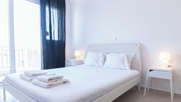 A modern hotel room with a large bed, white bedding, two bedside tables with lamps, and a view of the outdoors through a large window. - Gorgeous Apt w Magical View GC 3332-02 (Athens)
