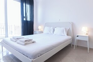 A modern hotel room with a large bed, white bedding, two bedside tables with lamps, and a view of the outdoors through a large window.