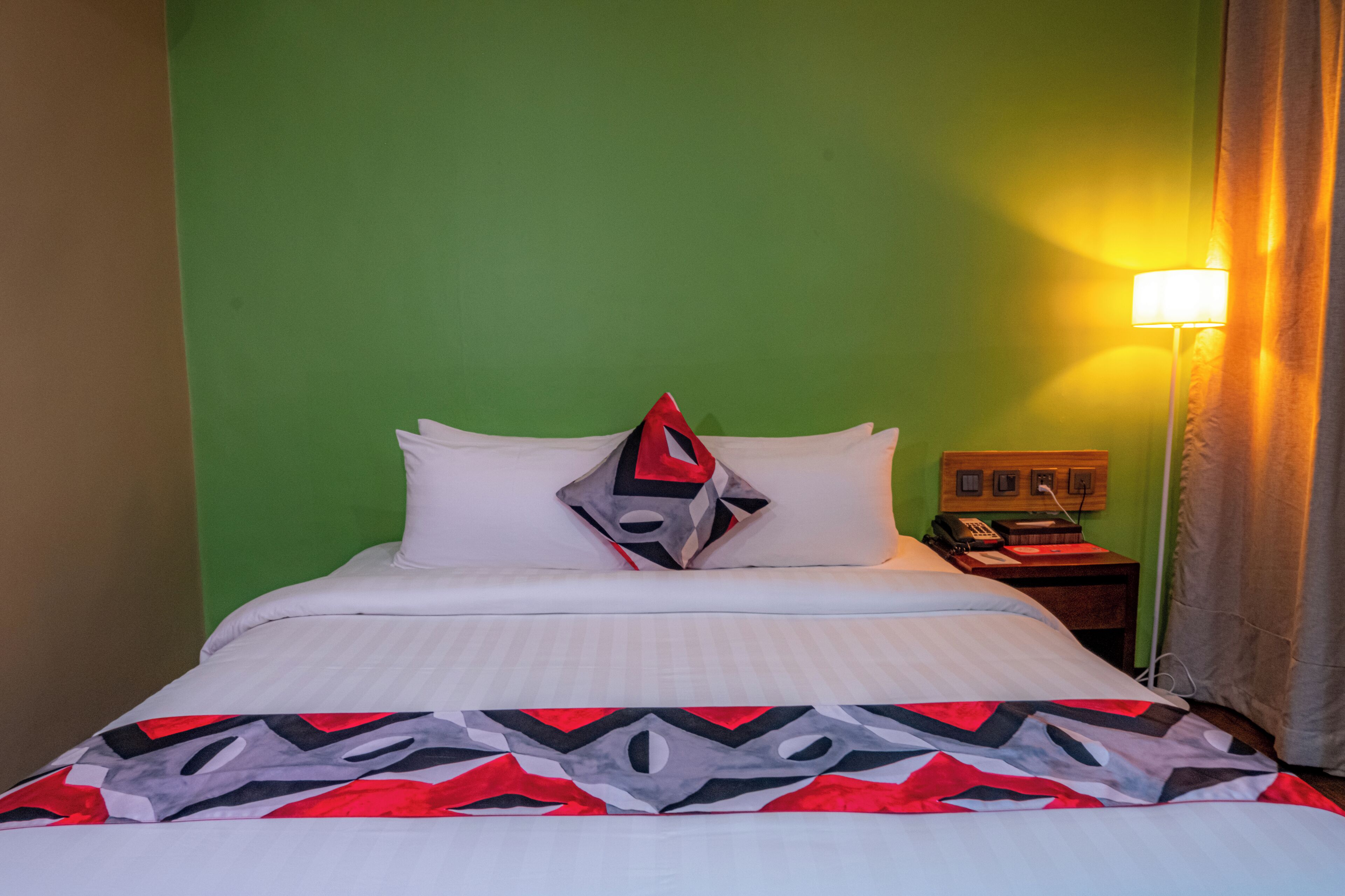 A hotel room with a green wall, a bed with white bedding, a patterned throw blanket, a nightstand with a lamp, and a telephone.