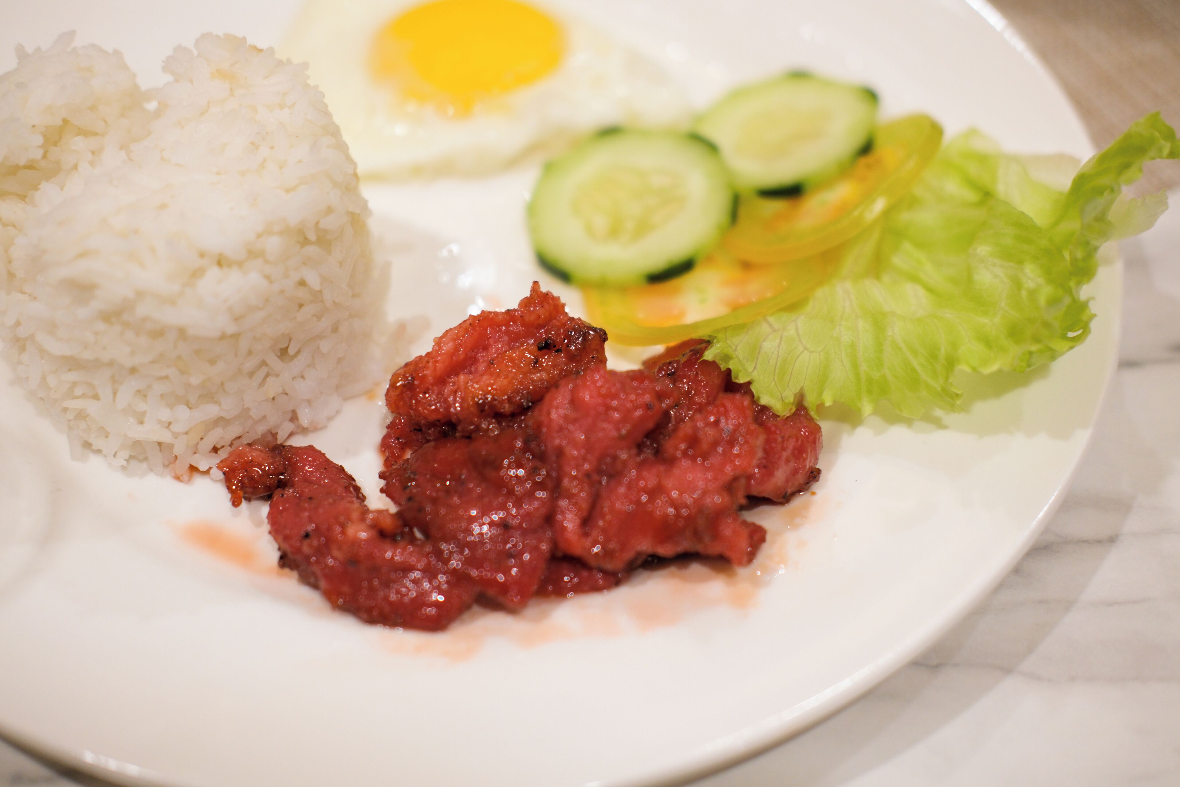 Un plato de comida con arroz, un huevo frito, rodajas de pepino, lechuga y un guiso de carne roja.