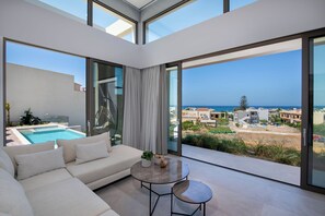 A modern living room with a sofa, a small table, and a view of a swimming pool and ocean. - Villa Manu Luxury Living (Chania)