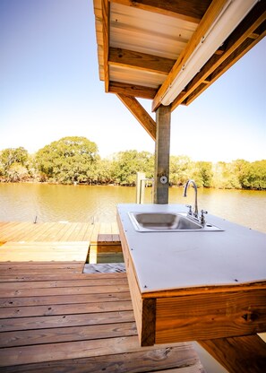 A wooden deck with a built-in sink and faucet, overlooking a calm body of water and lush greenery. - Sargent, Tx Spacious, Creekfront Home, Amazing Outdoor Space & 2 Fishing Piers. (Sargent)