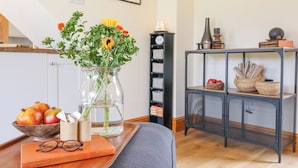 A cozy living room with a wooden coffee table holding a book, glasses, and a vase with flowers. A bookshelf with books and decorative items is in the background. - Brightside (Matlock)