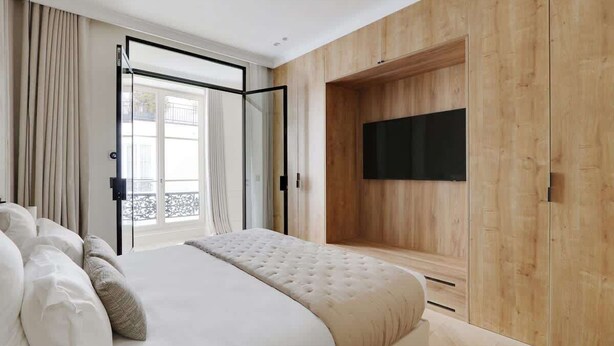 Un dormitorio con una cama grande, un televisor montado en una pared de madera y un ventanal corredero que da a un balcón exterior.