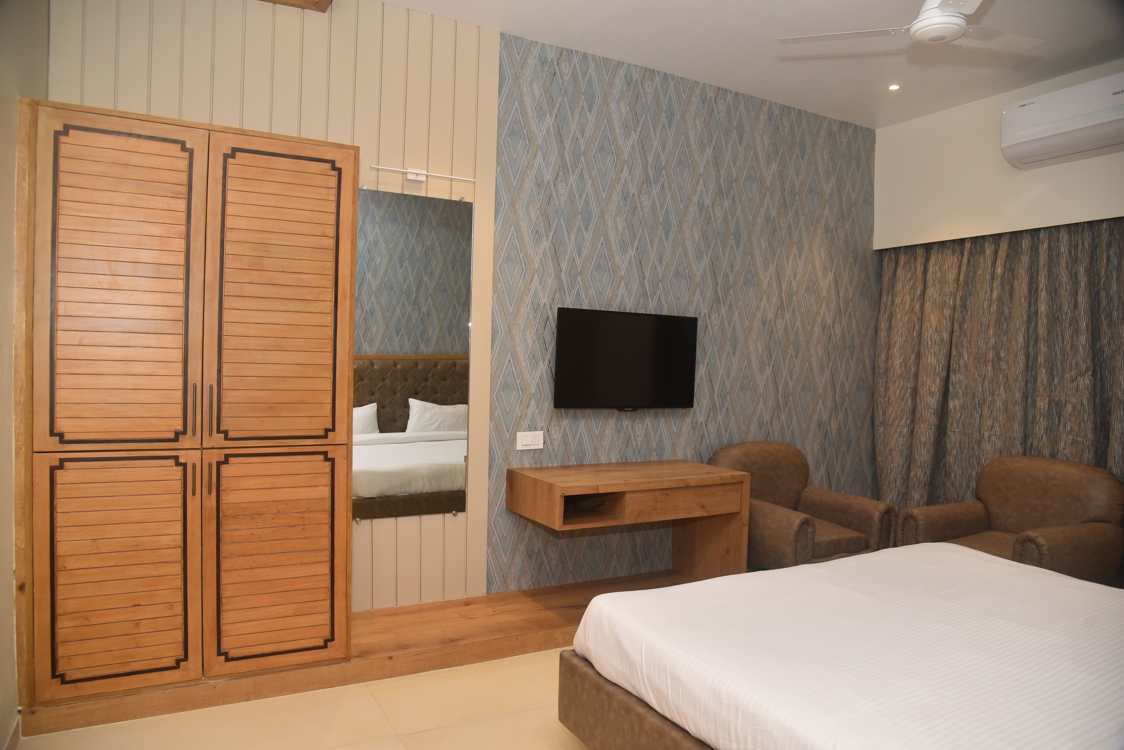 A bedroom with a bed, a TV mounted on the wall, a wooden wardrobe, and a desk with chairs.