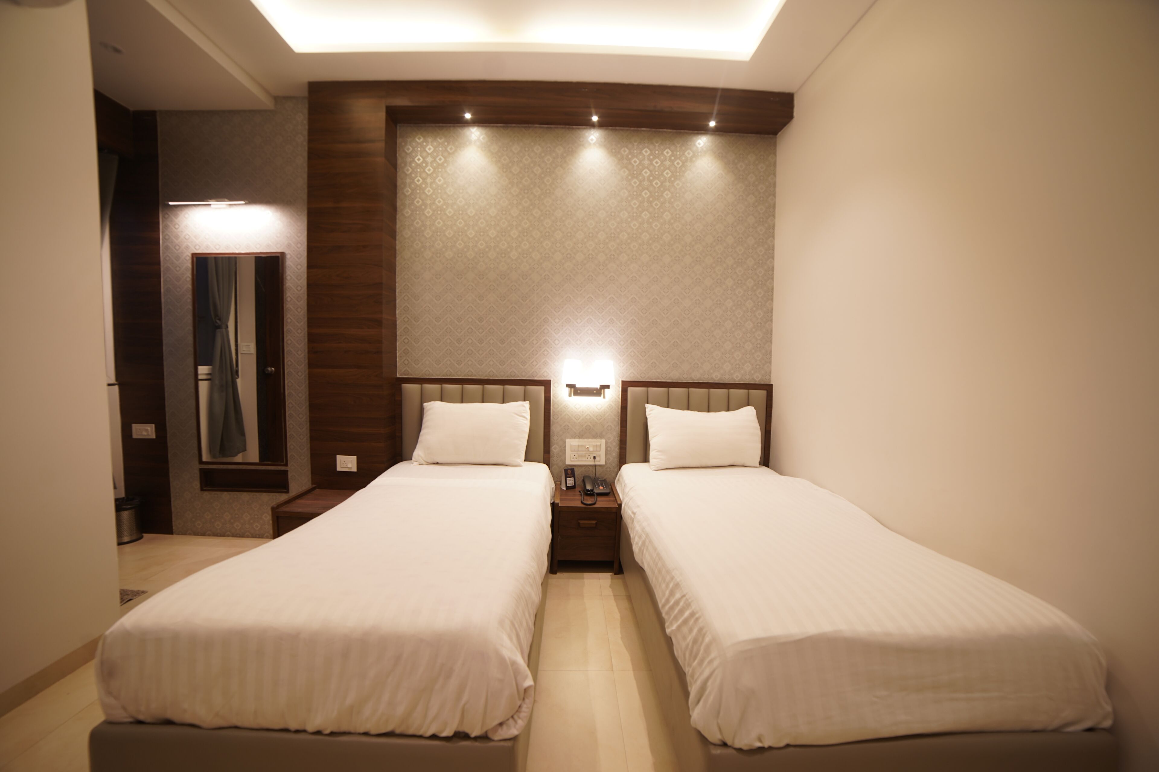 A hotel room with two single beds, a nightstand with a phone, a mirror, and a wall-mounted light fixture.
