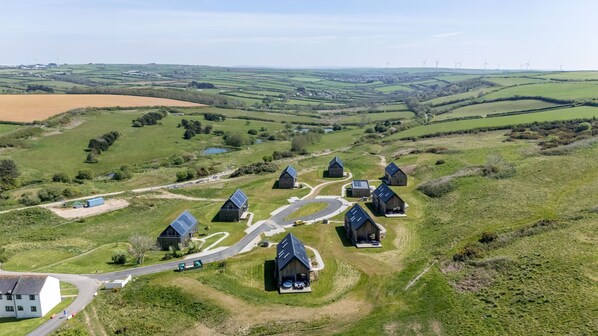 A rural area with several buildings, including a large white house and multiple dark-roofed structures, surrounded by green fields and wind turbines in the distance. - 4 The Vista, Willingcott Nr Woolacombe - Outdoor shower/ On-site pool / EV charger/ Ecohome (Woolacombe)