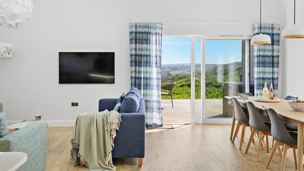 A modern living room with a sofa, a dining table with chairs, a TV mounted on the wall, and a large sliding glass door leading to an outdoor patio. - 4 The Vista, Willingcott Nr Woolacombe - Outdoor shower/ On-site pool / EV charger/ Ecohome (Woolacombe)