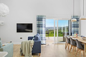 A modern living room with a sofa, a dining table with chairs, a TV mounted on the wall, and a large sliding glass door leading to an outdoor patio. - 4 The Vista, Willingcott Nr Woolacombe - Outdoor shower/ On-site pool / EV charger/ Ecohome (Woolacombe)