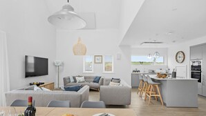 A modern kitchen with a central island, a dining area with a table set for two, and a living room with a sofa and TV. - 7 The Vista - Hot tub - Pet friendly - On-site pool - EV Charger - Ecohome (Woolacombe)