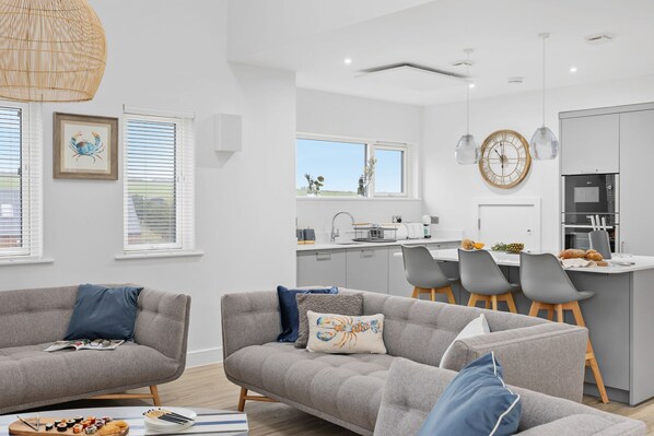 A modern living room with grey sofas, a dining area, and a kitchen with white cabinets.
