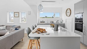 A modern kitchen with a central island, grey chairs, and a grey sofa in a bright room with large windows. - 7 The Vista - Hot tub - Pet friendly - On-site pool - EV Charger - Ecohome (Woolacombe)