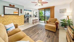 A spacious living area with a sofa, a chair, a kitchen in the background, and a balcony with a view. - Summer Deal Oceanfront Kona Condo with Sunset Views and Resort Pool (Kailua Kona)