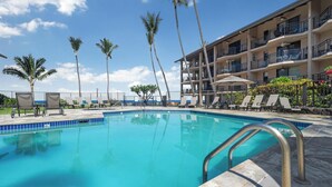 A swimming pool with lounge chairs and palm trees, surrounded by a multi-story building with balconies. - Summer Deal Oceanfront Kona Condo with Sunset Views and Resort Pool (Kailua Kona)