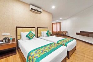 A hotel room with two beds, a desk with a chair, a small table with a kettle and water bottle, and a wall-mounted air conditioner.