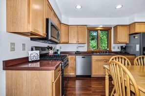 A modern kitchen with wooden cabinets, a stainless steel dishwasher, and a dining area with wooden chairs. - Stepaway Vacation Rentals - Hemlock House on Little St. Germain (St. Germain)