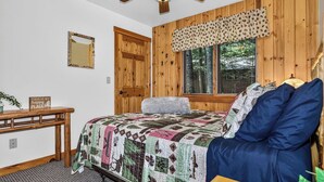 Une chambre avec un lambris en bois, un lit recouvert d’une courtepointe, un bureau avec un miroir et une fenêtre donnant sur des arbres.