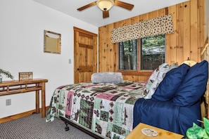 Un dormitorio con paneles de madera, una cama con colcha, un escritorio con espejo y una ventana con vistas a árboles.