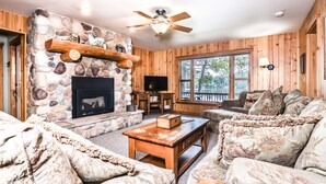 A cozy living room with a stone fireplace, wooden walls, a ceiling fan, and a large window overlooking a deck. - Stepaway Vacation Rentals - Hemlock House on Little St. Germain (St. Germain)