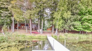 A wooden dock extending into a pond surrounded by trees, with a cabin in the background. - Stepaway Vacation Rentals - Hemlock House on Little St. Germain (St. Germain)