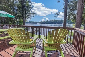 A deck with green Adirondack chairs and a table, overlooking a lake with trees and a clear sky.