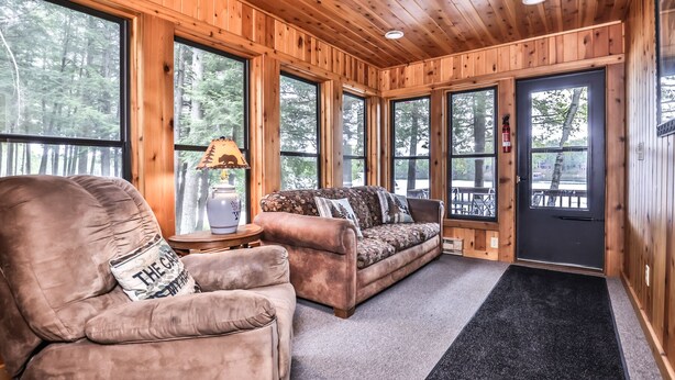 A cozy cabin interior with a plush armchair, a sofa, and a lamp with a bear design. Large windows offer views of a forested area. - Stepaway Vacation Rentals - Hemlock House on Little St. Germain (St. Germain)