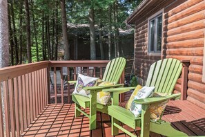 A wooden deck with two green Adirondack chairs and a small table, surrounded by a wooden fence and a log cabin. - Stepaway Vacation Rentals - Hemlock House on Little St. Germain (St. Germain)