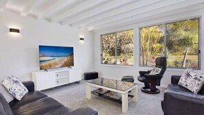 Uma sala de estar moderna com uma televisão grande, assentos confortáveis e amplas janelas com vista para árvores.