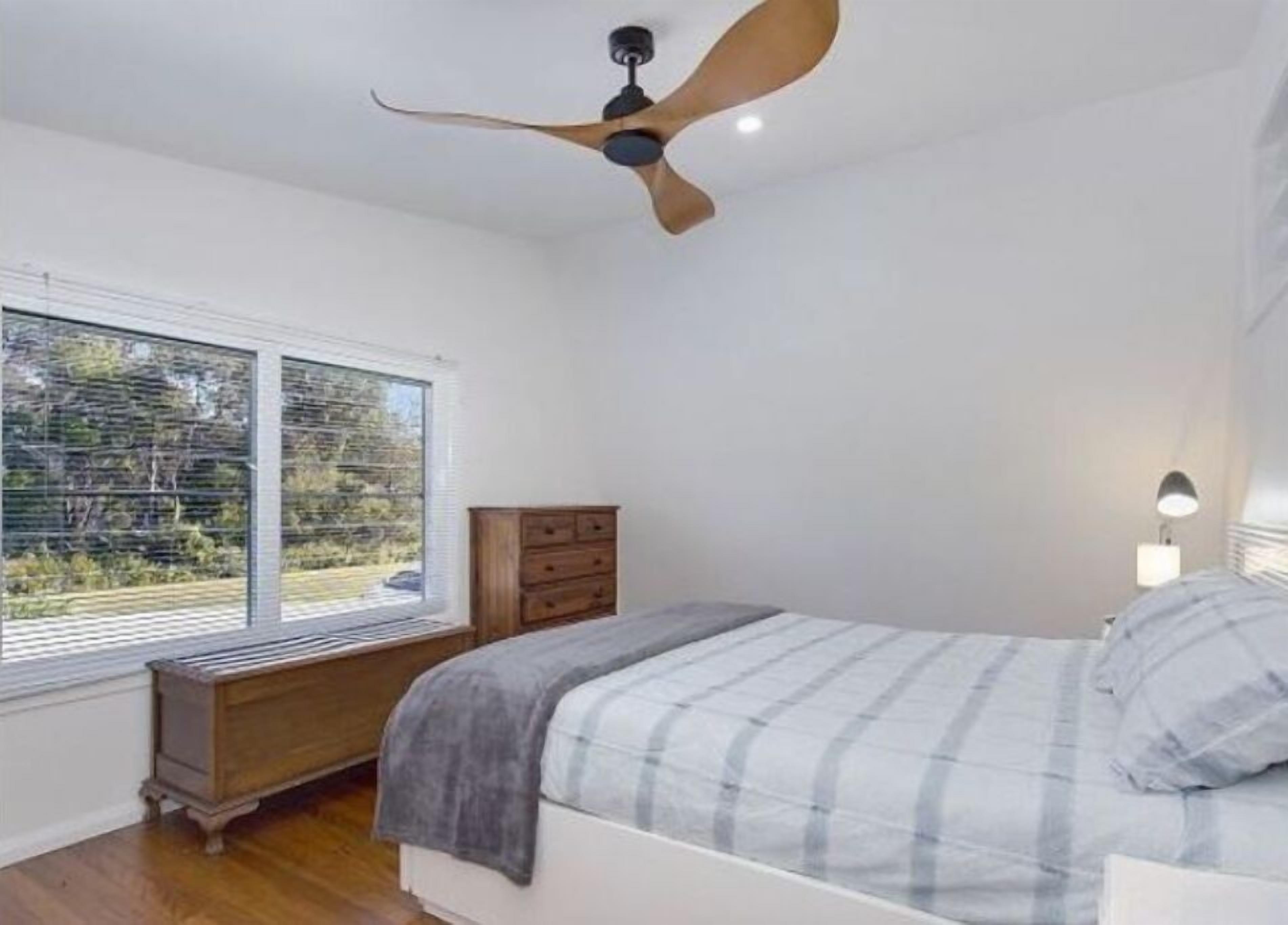 Een slaapkamer met een bed, een houten dressoir, een plafondventilator en een raam met uitzicht op bomen.
