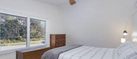 Een slaapkamer met een bed, een houten dressoir, een plafondventilator en een raam met uitzicht op bomen.