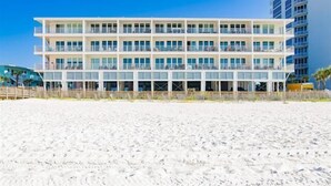 Un hôtel à plusieurs étages doté de balcons, situé sur une plage de sable fin, sous un ciel bleu azur.