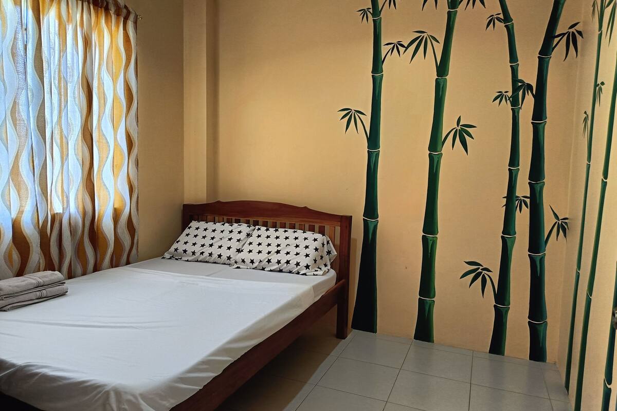 Un lit bien fait, avec un drap blanc et un oreiller à motifs, une tête de lit en bois et une fenêtre ornée de rideaux à motifs.