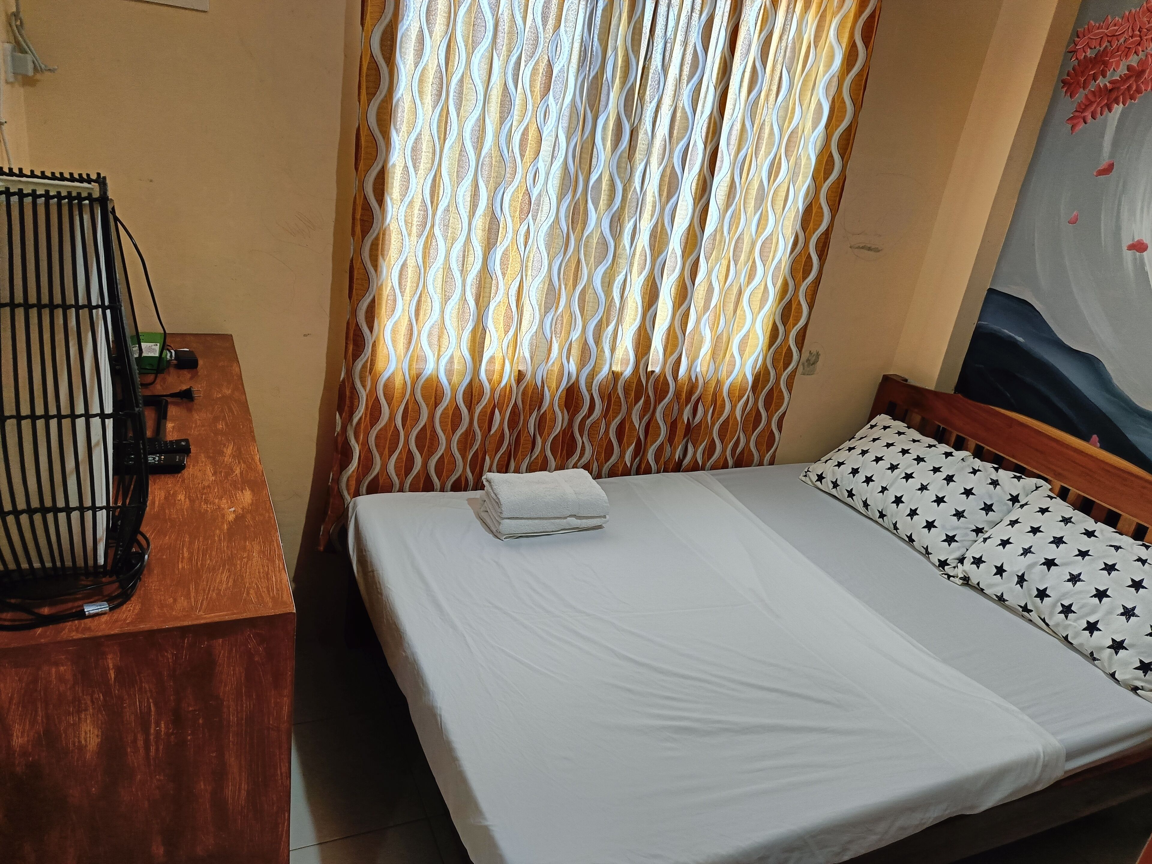 Un lit bien fait, avec un drap blanc et un oreiller orné d’un motif en étoile, une tête de lit en bois et un rideau à motifs ondulés.