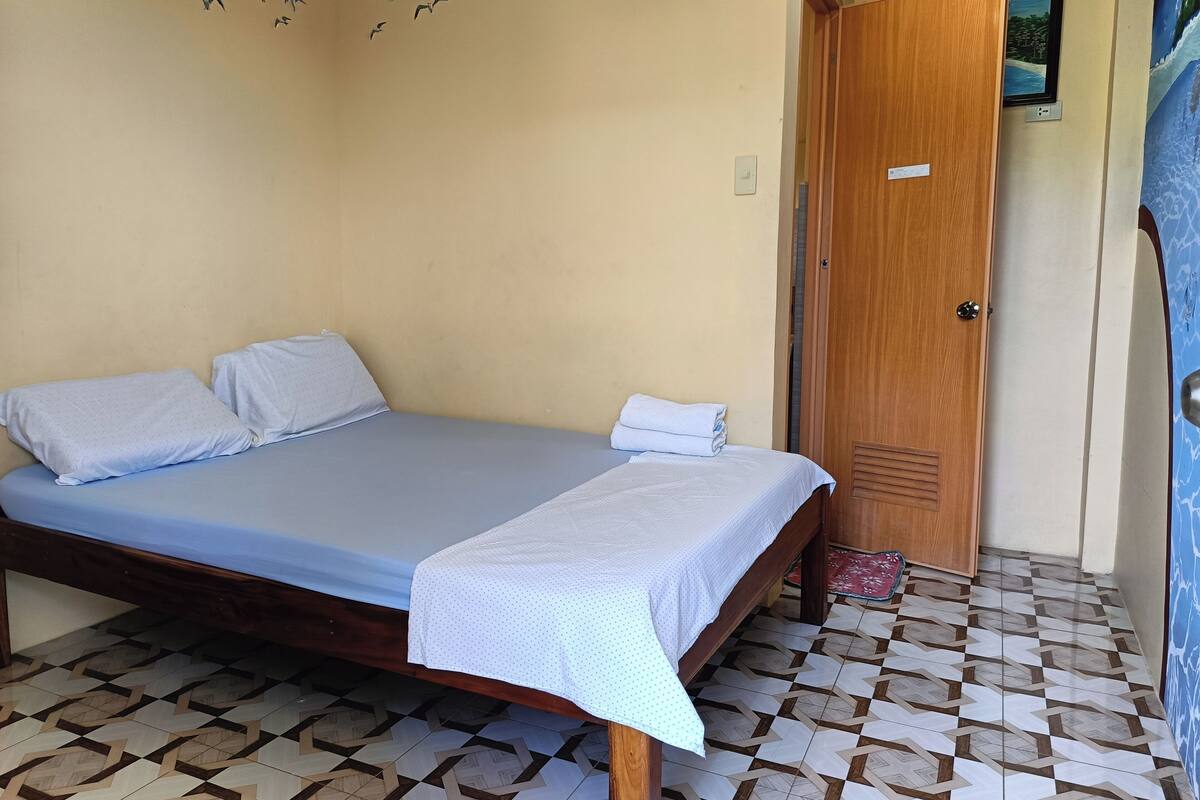 Un lit simple avec un matelas bleu et du linge de lit blanc, une porte en bois et des carreaux de sol à motifs.