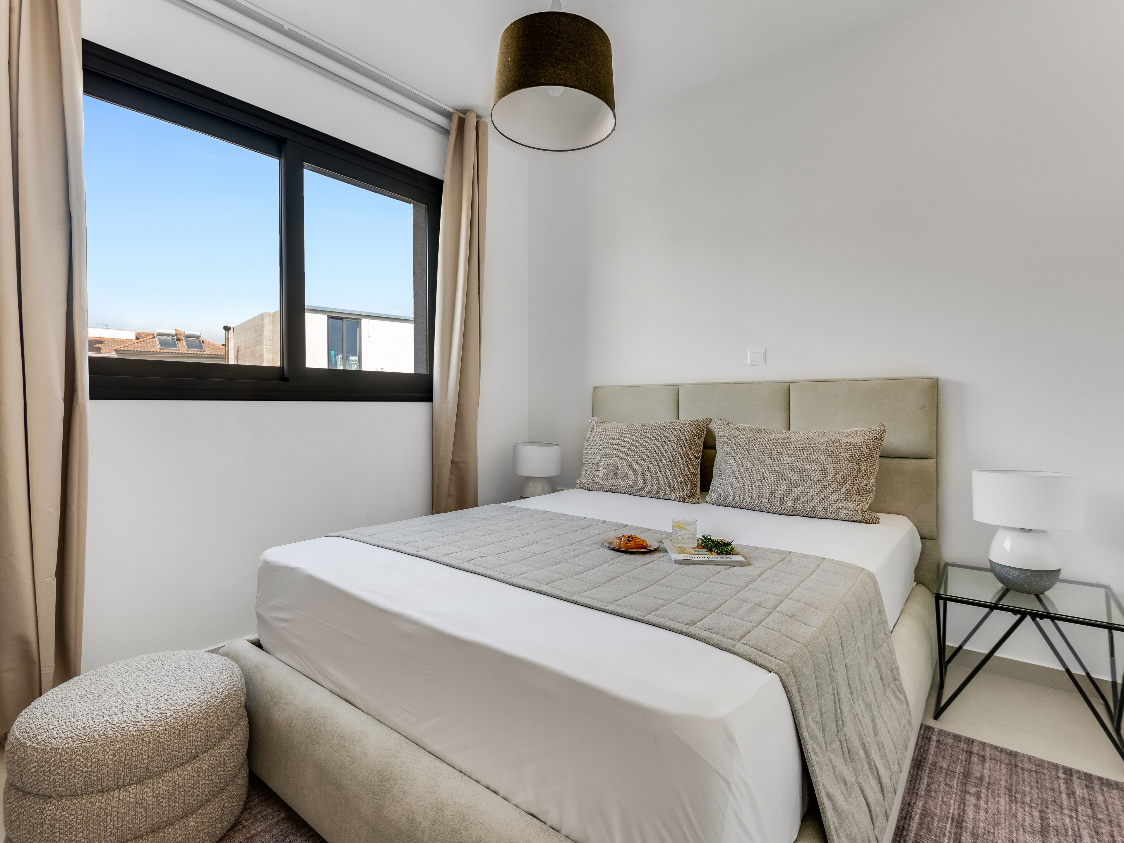 A hotel room with a bed, two bedside lamps, a window with curtains, a small table with a bowl, and a view of buildings outside.
