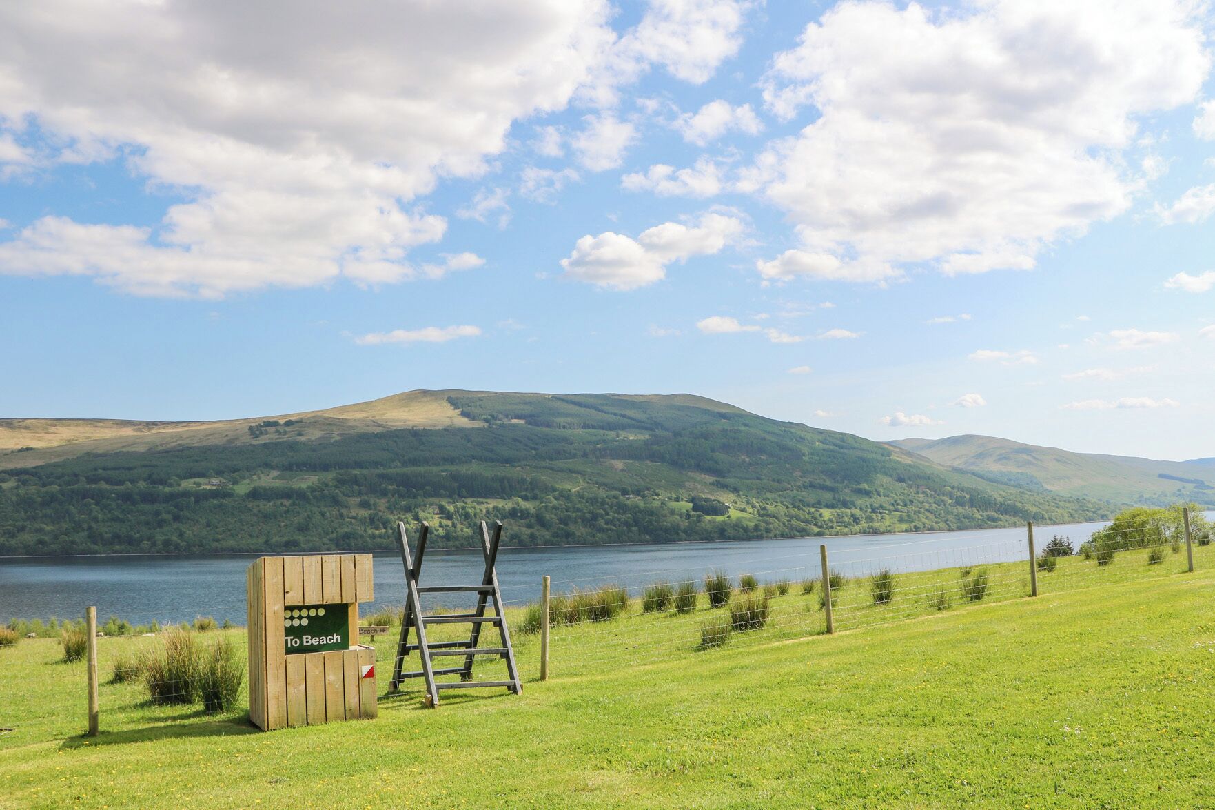 Une structure en bois portant l’inscription « Vers la plage », avec une échelle, un champ d’herbe et un lac, les montagnes se dressant à l’arrière-plan.