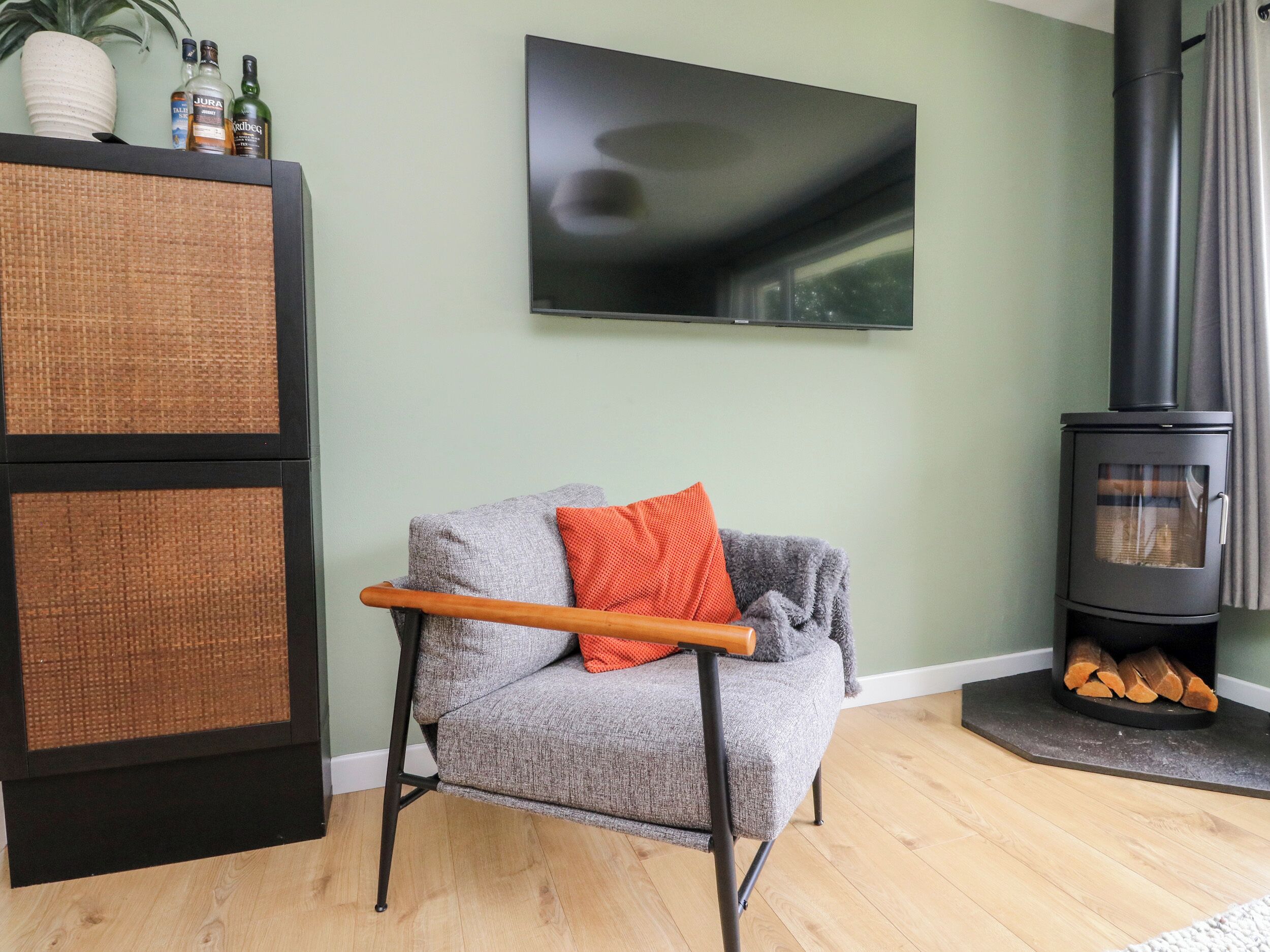 Un salón con un sillón gris, un televisor de pantalla plana, un hogar de leña negro y un aparador de mimbre.