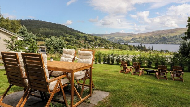 Un patio con sillas y una mesa de madera, con vistas a un paisaje pintoresco con colinas y un lago.