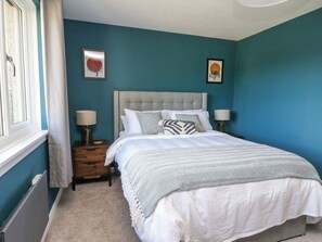 A bedroom with a grey upholstered headboard, a bed with white and patterned bedding, two bedside lamps, and two framed artworks on the wall. - Beech Cottage (Aberfeldy)