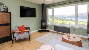 Una sala de estar con un sofá, un televisor, una chimenea y un ventanal con vistas a un lago y colinas.