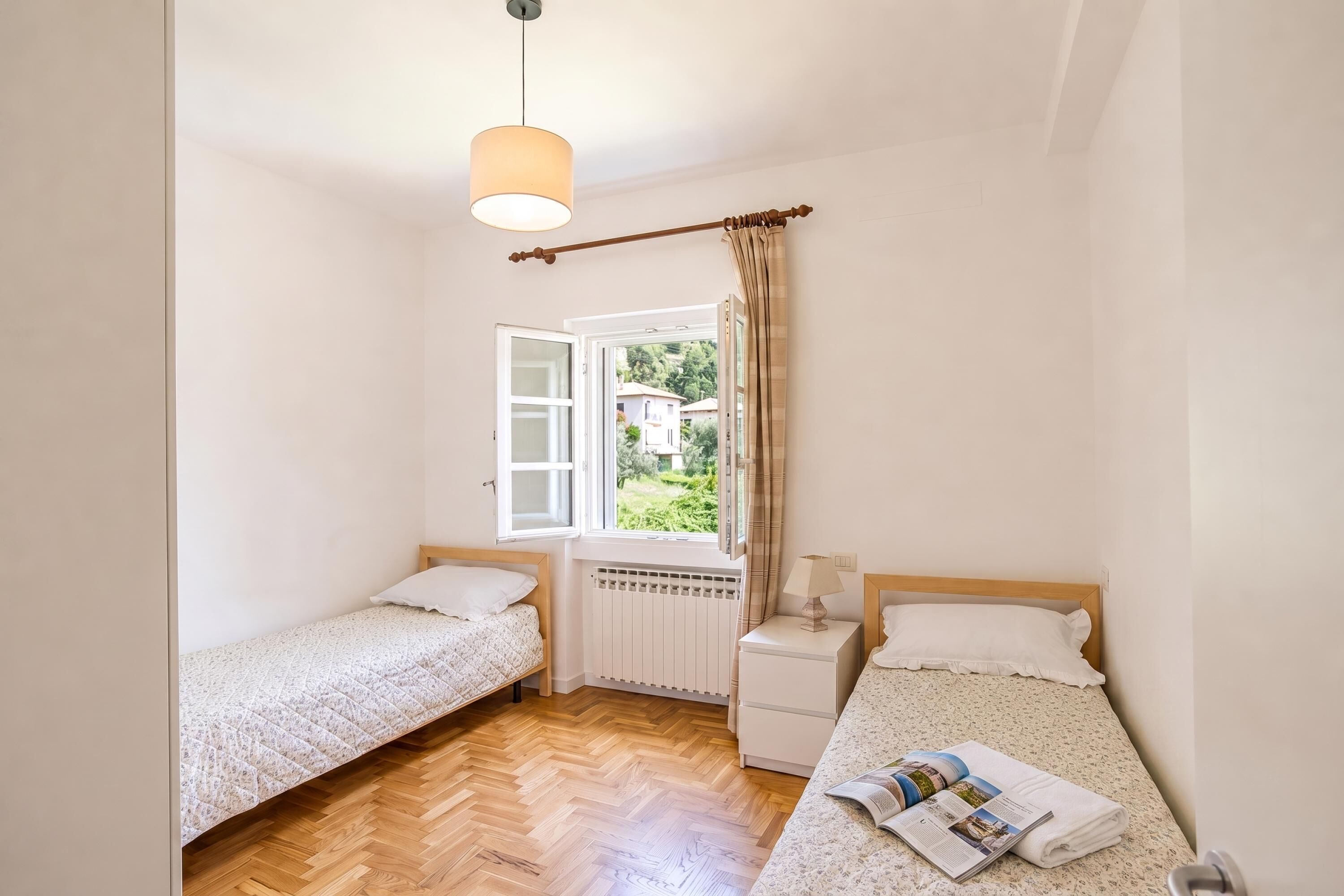A small, clean room with two single beds, a wooden floor, a window with curtains, and a radiator.