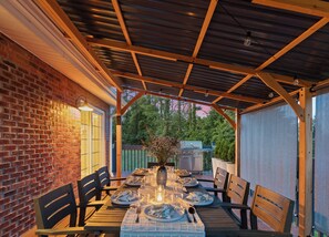 Une terrasse extérieure avec un mur en briques, des tables en bois disposées pour les repas et un espace couvert équipé d’un barbecue.