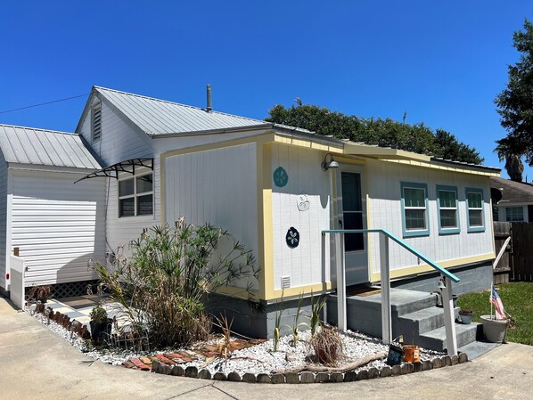 A small, single-story house with a white exterior and a yellow accent wall, a front porch, and a small garden area with plants and a flag.